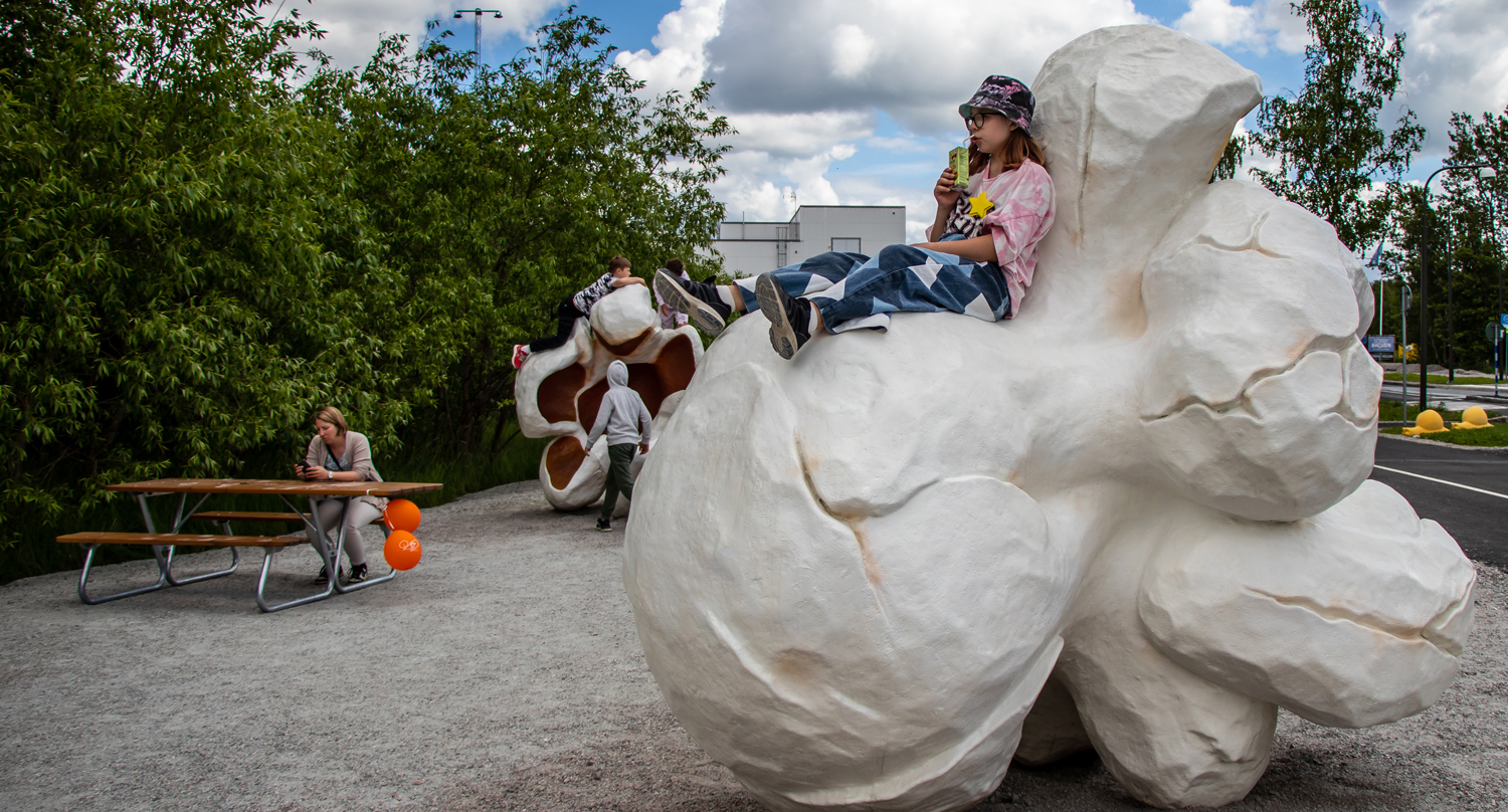 Vy över två jättepopcorn och en bänk. I förgrunden sitter en flicka på en popcorn och i bakgrunden leker pojkar vid ett annat popcorn. Vid bänken sitter en kvinna.
